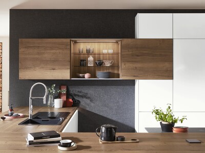 Perfektes Zusammenspiel von Farben und Licht. Die matte alpinweiße Küche und die dunkle Wand fassen Arbeitsplatte und Schränke aus Holzoptik optimal ein. Tolles Extra: Wandschrank mit Schiebetüren, der durch seine Innenbeleuchtung mehr Behaglichkeit bringt.